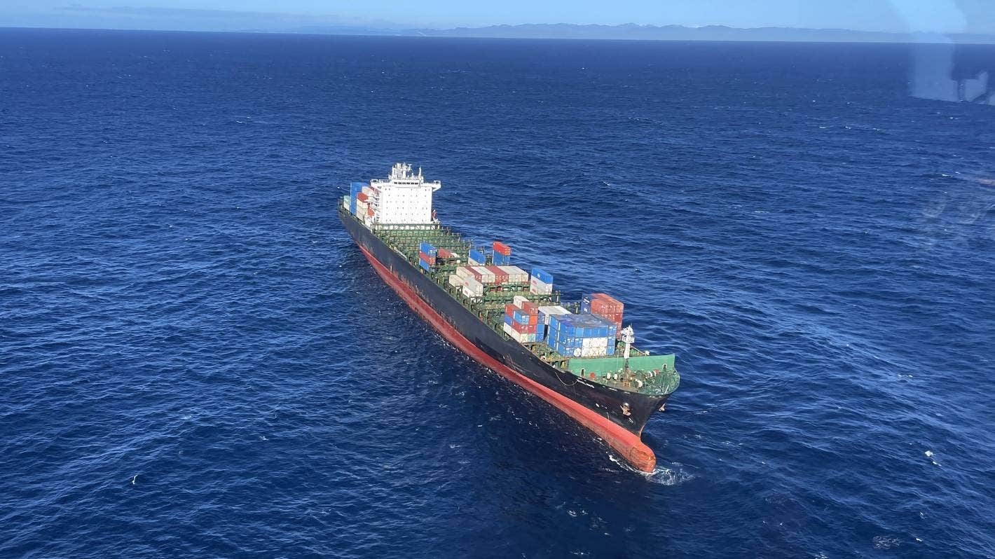 Aerial view of the container ship Shiling adrift in open ocean following a machinery failure during its second incident in 2023. The vessel is heavily laden with multi-coloured shipping containers stacked on deck. The bow is visibly trimmed low, and the ship appears to be listing slightly to port. Sea state is moderate under clear skies, with the New Zealand coastline faintly visible on the horizon. Image captured by the Nelson Marlborough Rescue Helicopter during a response operation