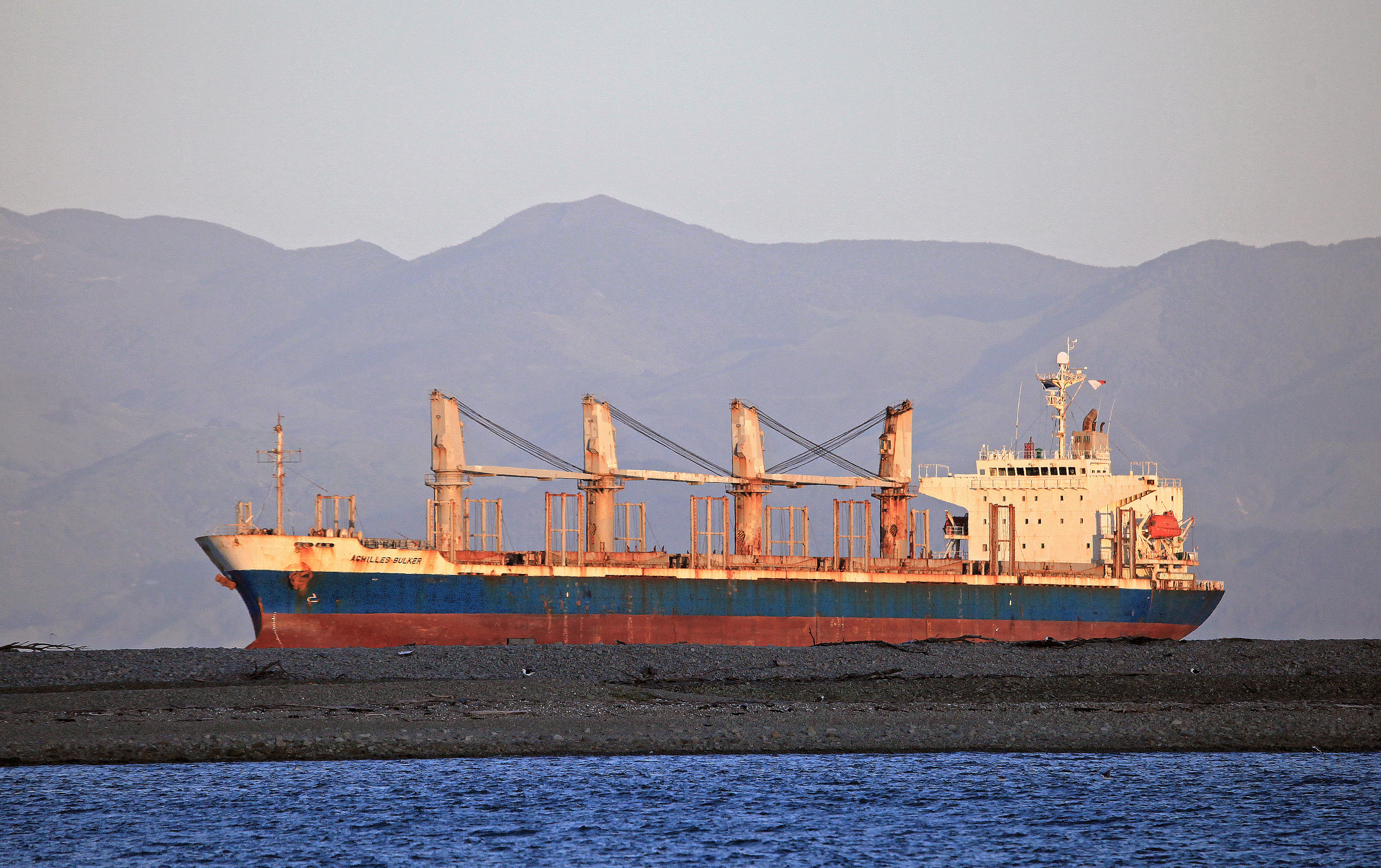  File photo of bulk carrier Achilles Bulker taken in 2022 near Nelson, New Zealand. The vessel is shown high out of the water, with anchor deployed, positioned behind a low-lying gravel or sandbar. Rust staining is visible along the hull, deck cranes, and superstructure. The ship has four cargo cranes and closed hatch covers, indicating a geared bulk carrier configuration. The vessel is lit by sunlight from a low angle, casting warm tones across the scene. Mountain ranges are visible in the background.