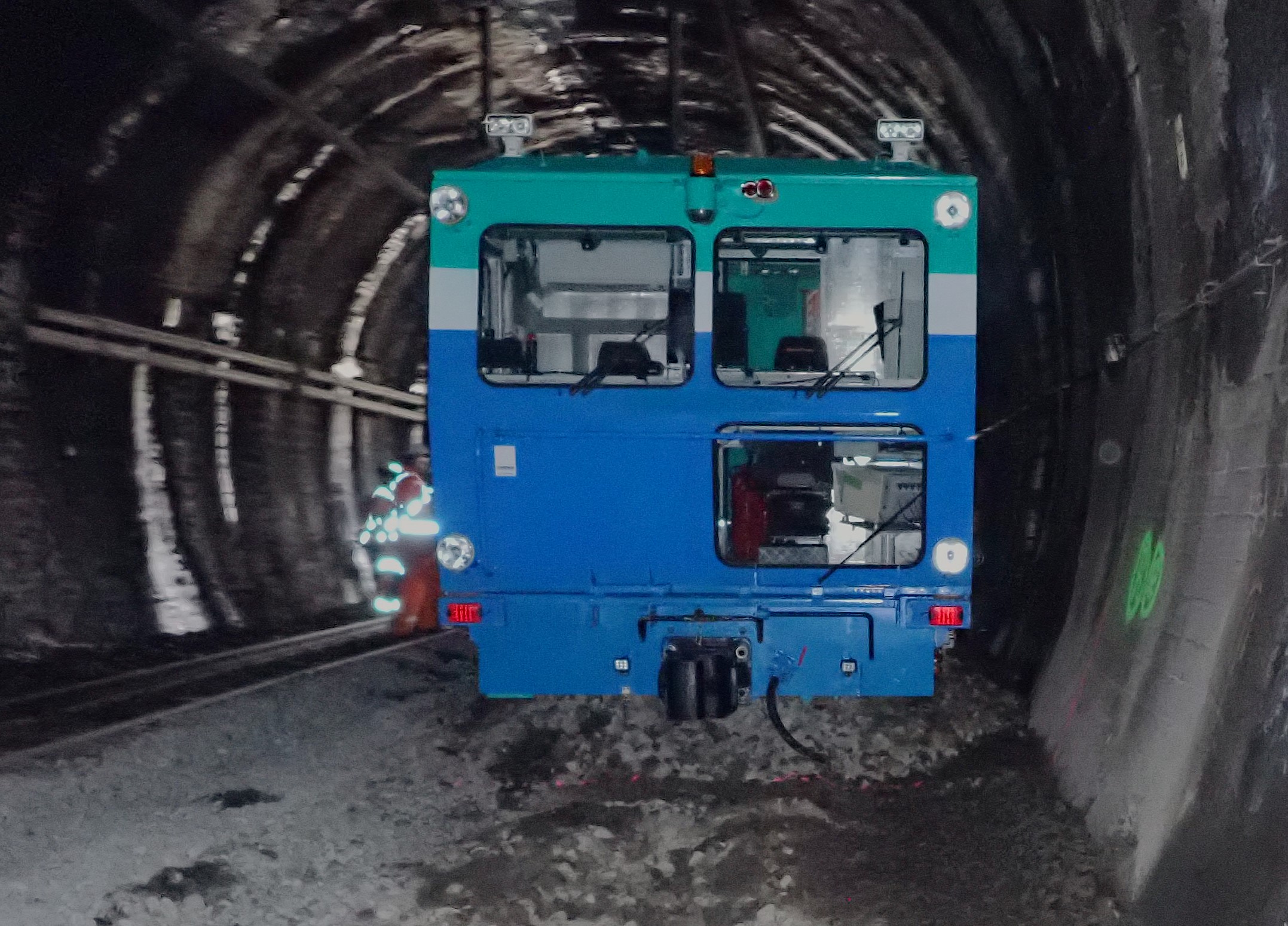 Front end view of a railway track tamper machine derailed inside a tunnel. The tamper has come to rest with a noticeable lateral tilt, indicating loss of alignment with the rails. A significant quantity of crushed stone ballast has been pushed forward and accumulated ahead of the machine, consistent with it having run approximately 16 metres off the rails through loose ballast. The image also shows the tunnel interior, tunnel wall markings, and components of the tamper's cab and underframe.