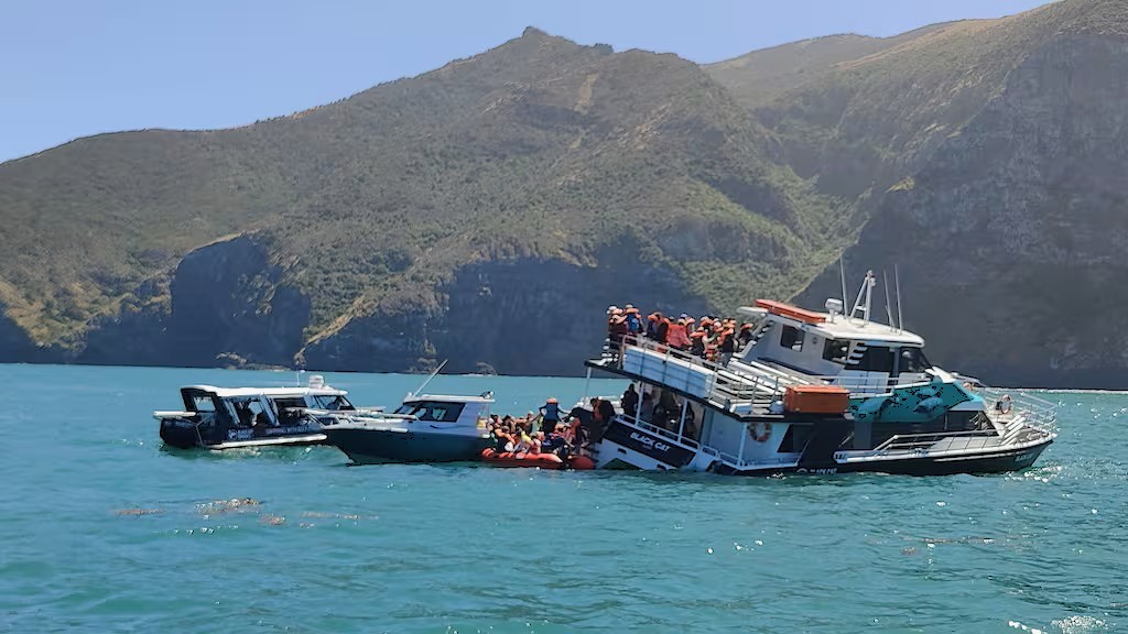 17-metre catamaran passenger ferry Black Cat photographed in Akaroa Harbour. No obvious major structural deformation above the waterline is visible. Vessel is listing to starboard by approximately 10° with starboard main-deck stern near to awash. Numerous people wearing orange lifejackets are visible on the main and upper deck on the port side, with some transferring on to one of three boats attending the stricken vessel's port side. Harbour headlands form the background. 