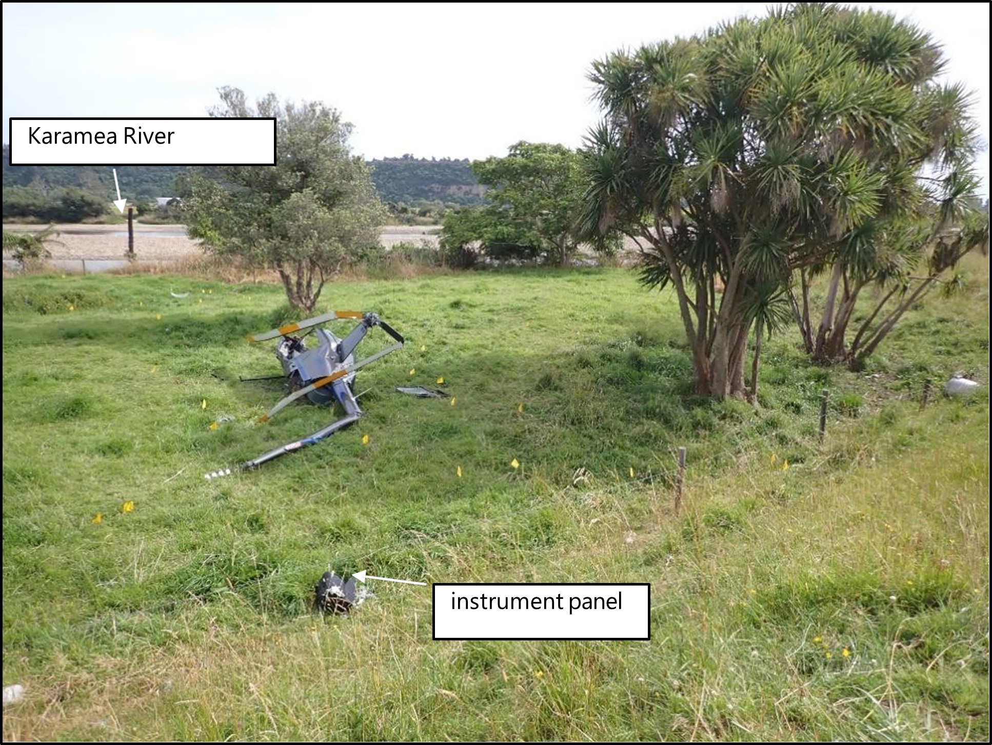 A crashed helicopter site near the Karamea River. Pieces of debris, including the detached instrument panel, are scattered around the crash site. The area is bordered by trees and shrubs.