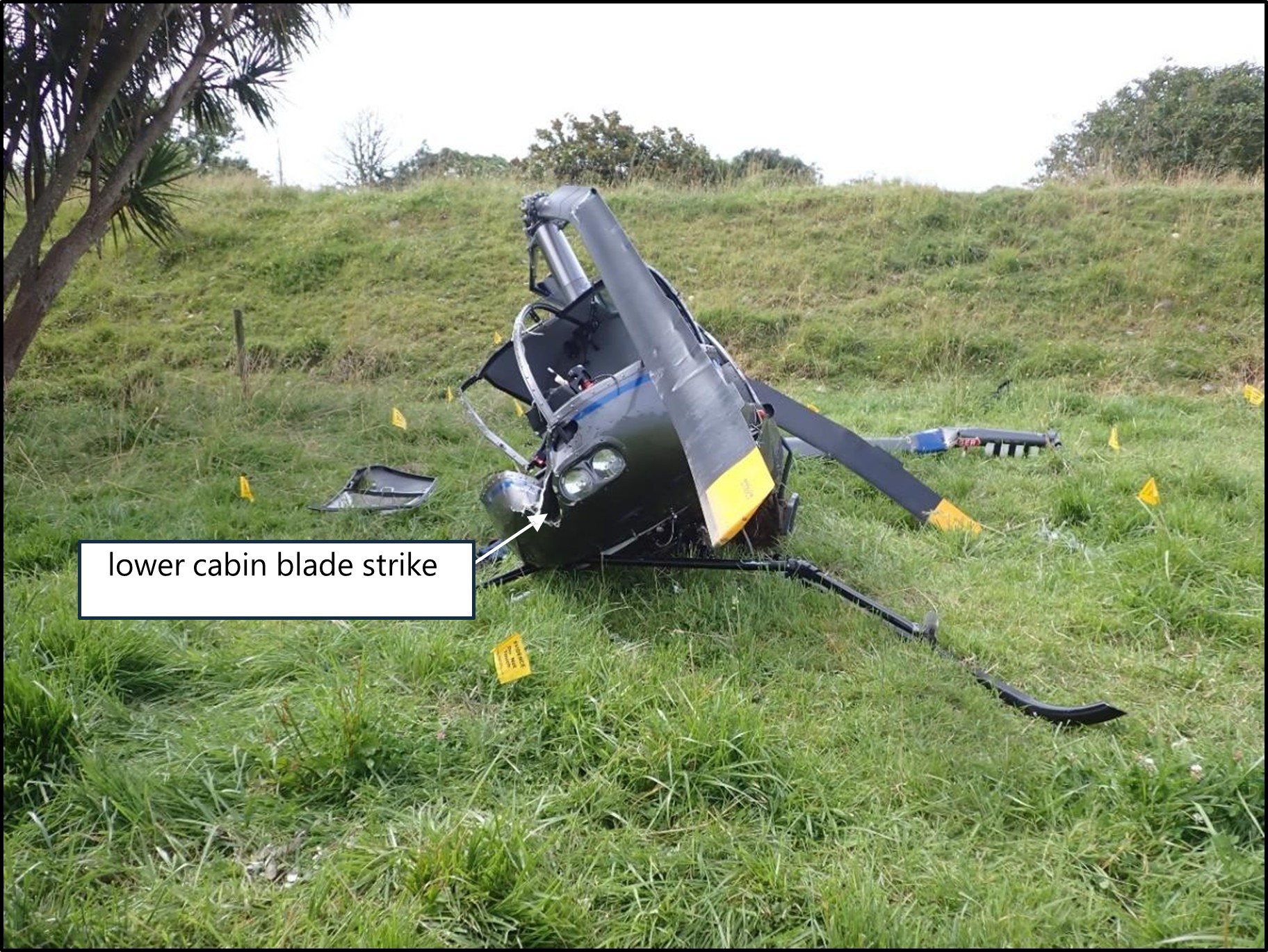 Photo of a crashed helicopter, showing significant damage to the lower cabin where a rotor blade struck. Pieces of debris are scattered nearby, with yellow evidence markers placed around the site.