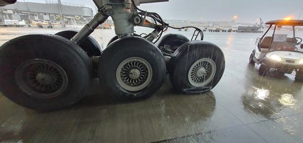 Close-up view of the right-hand main landing gear of a Boeing 777 aircraft on a wet airport apron during rainfall. The image shows six main gear tyres; the rear-most tyre (right side of image) is visibly deflated and compressed under load, with sidewall deformation. Other tyres appear fully inflated. Water runoff is visible on the gear structure and pooling on the concrete surface. A ground vehicle is present in the background to the right.