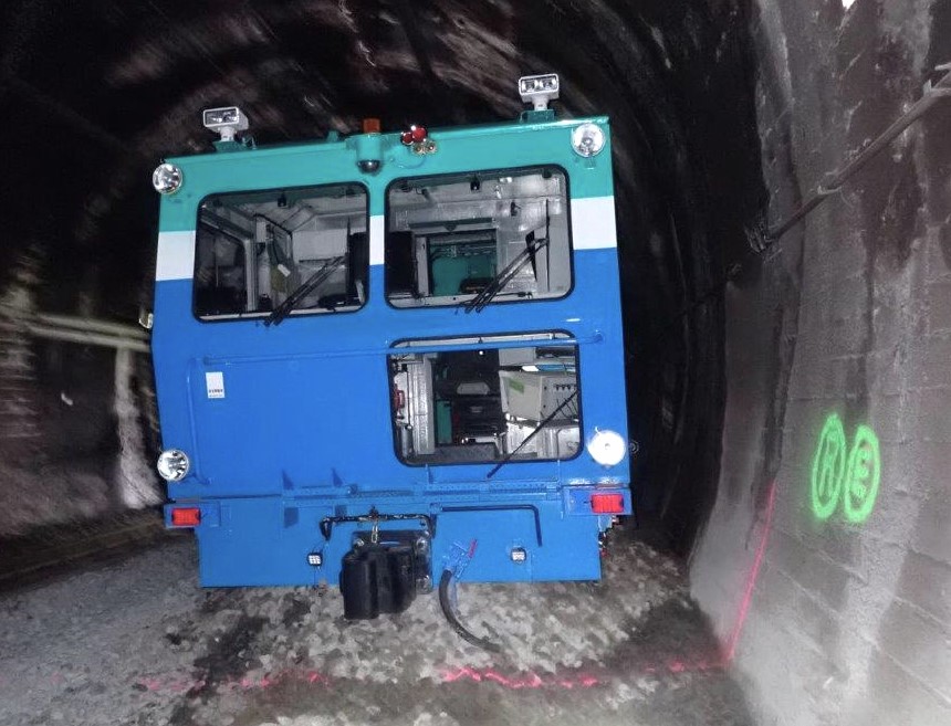Front end view of a railway track tamper machine derailed inside a tunnel. The tamper has come to rest with a noticeable lateral tilt, indicating loss of alignment with the rails. A significant quantity of crushed stone ballast has been pushed forward and accumulated ahead of the machine, consistent with it having run approximately 16 metres off the rails through loose ballast. The image also shows the tunnel interior, tunnel wall markings, and components of the tamper's cab and underframe.