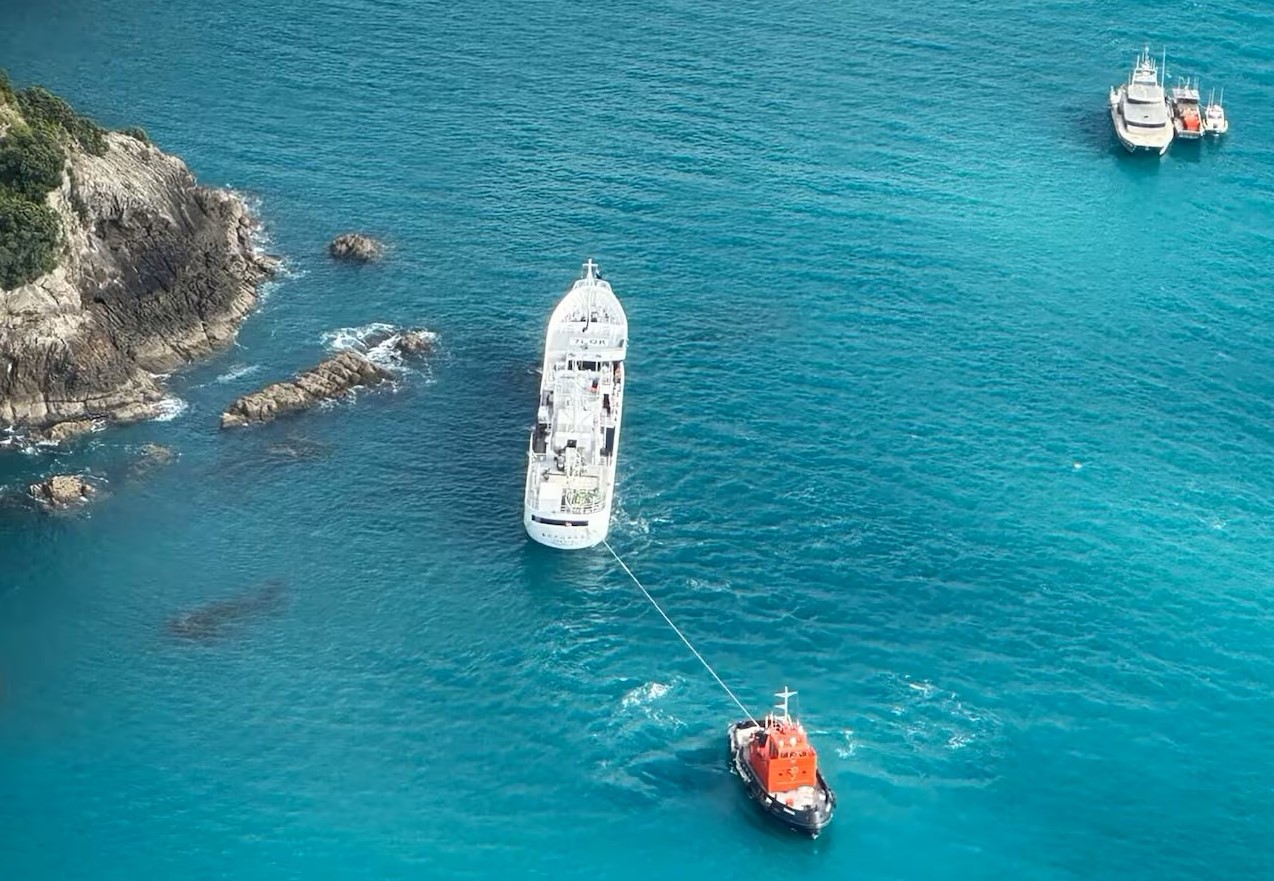 Aerial view of the Japanese longline fishing vessel Chokyo Maru aground on rocks near a steep coastline, with a harbour tug made fast to the vessel’s stern via a tow line. The tug is manoeuvring to refloat or reposition the grounded vessel. A group of support craft is visible in pontoon at anchor in deeper water some 60m off the starboard bow of the grounded vessel. The surrounding area features submerged hazards and clear turquoise water, indicating shallow depths and rocky outcrops