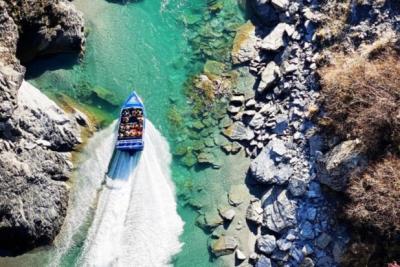 A blue jet boat speeds through a narrow, turquoise river surrounded by rocky cliffs and rugged terrain. The boat leaves a white water trail as it navigates the clear, shallow waters, carrying passengers who are tightly seated and facing forward. The scene is captured from above, showcasing the contrast between the vibrant water and the gray stone landscape.