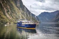 The Fiordland Navigator. File shot supplied. A large blue and yellow passenger vessel named 'Fiordland Navigator' is anchored on calm water in a mountain fjord. The vessel has three tall masts, each with a furled jib sail for optional wind-powered sailing. It features three decks with rows of windows, and an open front viewing platform. In the background, a small craft moves across the water. The fjord is surrounded by steep, forested mountains under a cloudy sky.