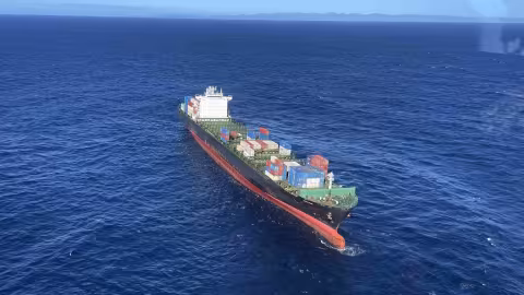 Aerial view of the container ship Shiling adrift in open ocean following a machinery failure during its second incident in 2023. The vessel is heavily laden with multi-coloured shipping containers stacked on deck. The bow is visibly trimmed low, and the ship appears to be listing slightly to port. Sea state is moderate under clear skies, with the New Zealand coastline faintly visible on the horizon. Image captured by the Nelson Marlborough Rescue Helicopter during a response operation