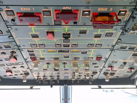 Photo taken in the cockpit of an A320 from behind the pilot seats, looking forward, showing the overhead control panel. Nearest the viewer at the top of the photo is the fire control panel for Engine 1, auxiliary power unit and Engine 2. The push-release fire control switch for Engine 2 is in the ‘popped-out’ activated position. Beyond the fire panel are panels for hydraulics, fuel, electrical, with illuminated indicators, guarded switches, rotary selectors, and other controls, standard A320 layout.