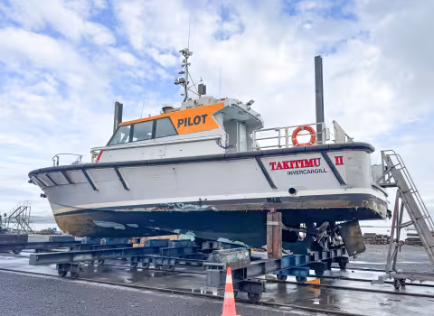 Port-side view of the pilot launch Takitimu II, lifted out of the water and supported on a wheeled slipway cradle. The vessel’s hull, wheelhouse marked ‘PILOT’, and superstructure are visible, along with navigation and communications antennas mounted on the mast. The propeller, rudder, and lower hull are exposed, showing damage to the hull, propellers and steering gear from grinding on rocks after the grounding. The vessel is positioned ashore in a shipyard environment under partly cloudy conditions