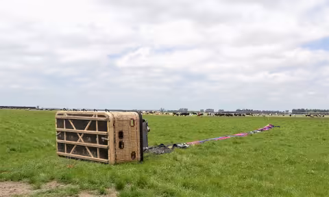 photo depicts balloon basket lying on its side in a cow paddock. The deflated balloon envelope is laid out partially folded across the grass 