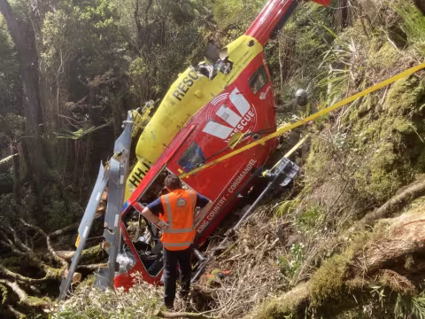 The wreckage of a crashed BK117 rescue helicopter sits upright in dense bush on a steep slope. The helicopter has a red fuselage with 'RESCUE' and 'King Country – Coromandel' markings, and a yellow tail boom. the main rotor blades are torn up, ripped and distorted from slashing through the trees during the crash sequence. A man in a high-visibility vest bearing the TAIC logo stands with his back to the viewer, looking at the wreck. Another person in rescue gear is partially visible among the vegetation.