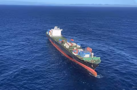 Aerial view of the container ship Shiling adrift in open ocean following a machinery failure during its second incident in 2023. The vessel is heavily laden with multi-coloured shipping containers stacked on deck. The bow is visibly trimmed low, and the ship appears to be listing slightly to port. Sea state is moderate under clear skies, with the New Zealand coastline faintly visible on the horizon. Image captured by the Nelson Marlborough Rescue Helicopter during a response operation