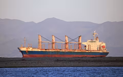  File photo of bulk carrier Achilles Bulker taken in 2022 near Nelson, New Zealand. The vessel is shown high out of the water, with anchor deployed, positioned behind a low-lying gravel or sandbar. Rust staining is visible along the hull, deck cranes, and superstructure. The ship has four cargo cranes and closed hatch covers, indicating a geared bulk carrier configuration. The vessel is lit by sunlight from a low angle, casting warm tones across the scene. Mountain ranges are visible in the background.