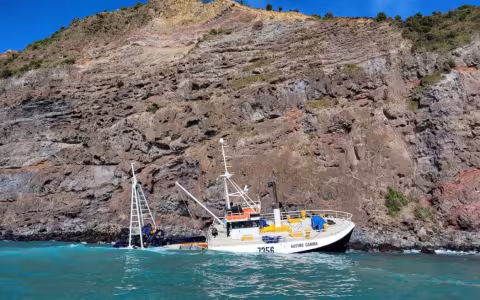 A fishing vessel named Austro Carina is partially submerged and leaning against a rocky and high cliff. The stern of the vessel is in the water, while the bow remains above the surface. The surrounding water is turquoise, and the cliff has layers of brown and reddish rock with sparse green vegetation. The sky is clear and blue.